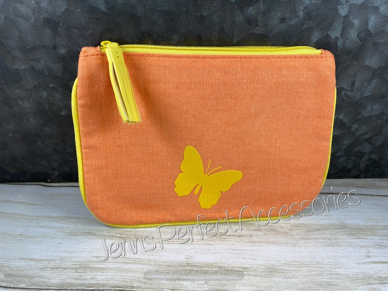 May include: Orange canvas zippered pouch with a yellow butterfly applique. The pouch has a yellow zipper and trim, and a yellow tassel on the zipper pull. The text "Jenns Perfect Accessories" is printed on the bottom.