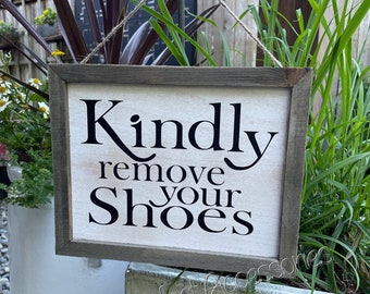 May include: A rectangular wooden sign with a gray frame and the text "Kindly remove your Shoes" in black. The sign is hanging by a twine string. The background includes greenery and a white planter.