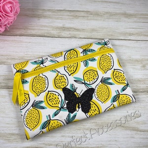 May include: A rectangular zippered pouch with a white background and a pattern of yellow lemons with green leaves. A black glitter butterfly is centered on the pouch. The zipper and pull are yellow. The text "Jenns Perfect Accessories" is printed on the bottom.