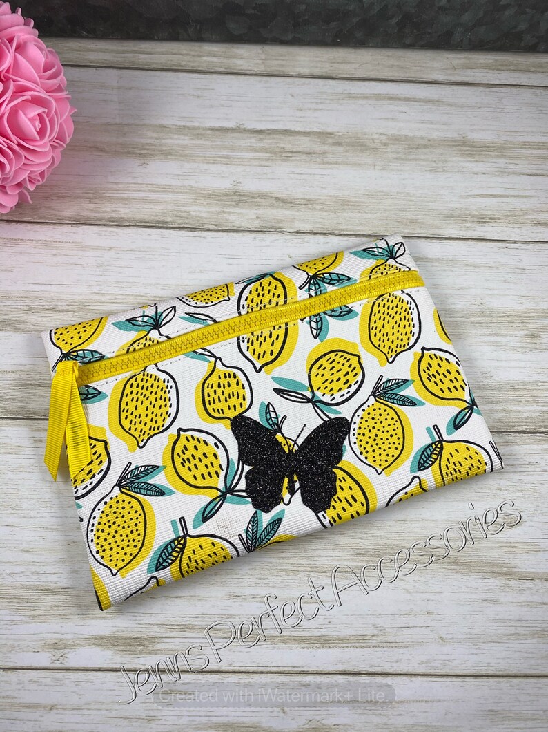 May include: A rectangular zippered pouch with a white background and a pattern of yellow lemons with green leaves. A black glitter butterfly is centered on the pouch. The zipper and pull are yellow. The text "Jenns Perfect Accessories" is printed on the bottom.