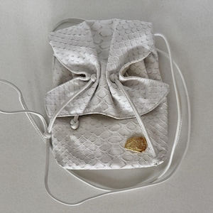 May include: A white drawstring bag featuring a textured snakeskin pattern. The bag has a folded top flap secured by thin white cords and a small, irregular gold-toned metal emblem attached to the lower front section of the exterior surface.