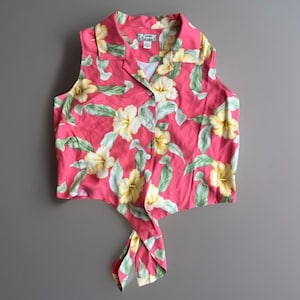 May include: A sleeveless button-down shirt in a vibrant pink hue featuring a repeating pattern of pale yellow hibiscus flowers and light green leaves. The garment has a notched collar and a front hem that ties into a knot at the center.