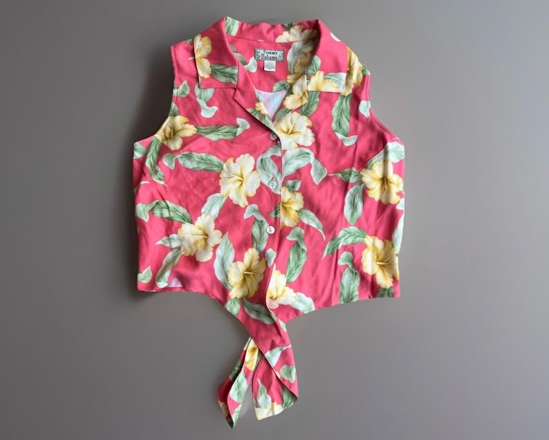 May include: A sleeveless button-down shirt in a vibrant pink hue featuring a repeating pattern of pale yellow hibiscus flowers and light green leaves. The garment has a notched collar and a front hem that ties into a knot at the center.