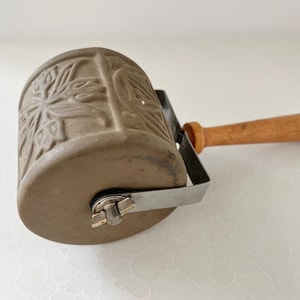 May include: A cylindrical clay roller tool featuring an embossed floral pattern on its surface. The roller is attached to a metal frame with a wing nut and includes a smooth, light brown wooden handle extending from the center of the frame.