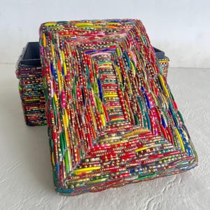 May include: A rectangular box with a lid, covered in a mosaic of colorful beads. The beads are arranged in a geometric pattern, with gold accents. The box is open, revealing a dark interior.