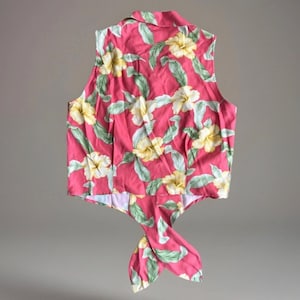 May include: A sleeveless button-down shirt in a vibrant pink hue, featuring a repeating pattern of pale yellow hibiscus flowers and light green leaves. The garment has a pointed collar and a front hem that ties into a knot at the center waist.