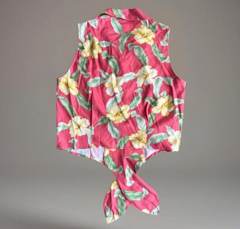 May include: A sleeveless button-down shirt in a vibrant pink hue, featuring a repeating pattern of pale yellow hibiscus flowers and light green leaves. The garment has a pointed collar and a front hem that ties into a knot at the center waist.