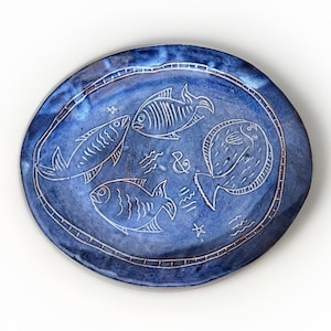 May include: An oval ceramic platter with a deep blue glaze featuring white etched illustrations of three fish, a small star, and an ampersand symbol. The rim is decorated with a thin, etched border line that follows the oval shape of the platter.