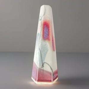 May include: A tall, tapered, multi-faceted ceramic sculpture with a white base glaze. The surface features abstract, blended patterns in shades of deep red, soft purple, and muted blue-grey, creating a smooth, gradient effect across the object.