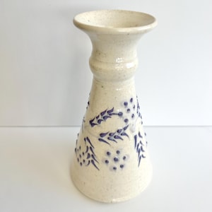May include: Cream-colored ceramic candlestick holder with a flared top and base. The body features a textured surface and is decorated with dark blue floral patterns. The candlestick is approximately 8 inches tall.