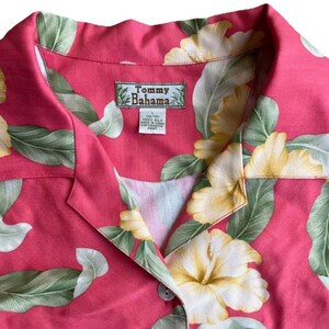 May include: A pink short-sleeved shirt featuring a tropical print of large yellow hibiscus flowers and green leaves. The shirt has a notched collar and a Tommy Bahama brand label sewn into the inner neckline, indicating 100% silk material.