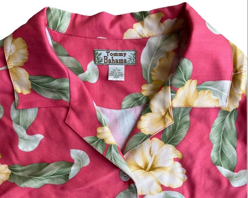 May include: A pink short-sleeved shirt featuring a tropical print of large yellow hibiscus flowers and green leaves. The shirt has a notched collar and a Tommy Bahama brand label sewn into the inner neckline, indicating 100% silk material.