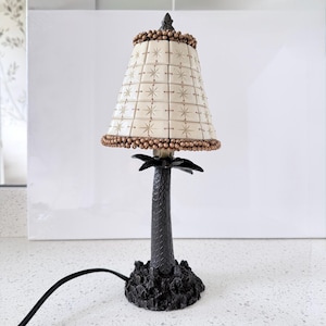 May include: A table lamp featuring a dark brown, textured palm tree base and a conical lampshade. The shade is constructed from cream-colored square tiles, each etched with a starburst pattern, and is trimmed with small brown beads at the top and bottom.