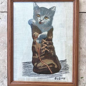 May include: Embroidered artwork of a gray cat wearing a brown boot. The cat has yellow eyes and is sitting on a black and white striped surface. The artwork is in a brown wooden frame. The name "SUSAN" is embroidered on the bottom right.