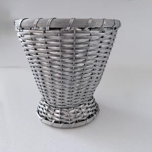 May include: A silver-colored metallic basket with a woven pattern, featuring a flared top rim and a circular base. The structure is composed of thin, interconnected metal strips that create a consistent, repeating lattice design throughout the body.