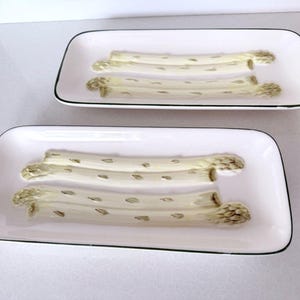 May include: Two rectangular white ceramic serving platters with rounded corners and a thin black rim. Each platter features a raised, three-dimensional molded design of two white asparagus stalks with light green tips centered on the surface.
