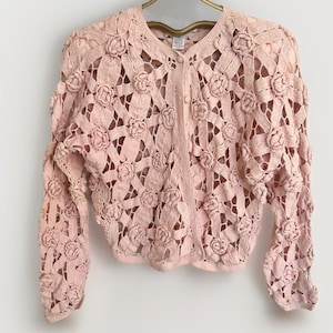 May include: A pale pink long-sleeve cardigan featuring an intricate openwork crochet pattern with raised floral motifs throughout. The garment has a round neckline, a front button closure with matching buttons, and a cropped, structured silhouette.