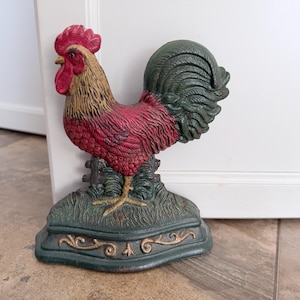 May include: A cast iron doorstop shaped like a rooster with a red comb and wattle, a tan neck, a red body, and a dark green tail. The rooster stands on a green base with a textured grass pattern and gold-colored decorative scrollwork details.