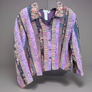May include: A button-front jacket featuring a patchwork design of vertical stripes in shades of purple, black, and multi-colored floral embroidery. The garment has a pointed collar, long sleeves, and a purple interior lining with a Coldwater Creek label.