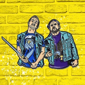 May include: Two enamel pins featuring cartoon characters wearing denim jackets and eagle graphic t-shirts. One character holds a sword and the other holds a gun.