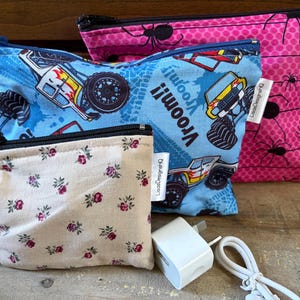 May include: Three zippered pouches of varying sizes. The smallest is beige with a small red and green floral pattern. The medium pouch is blue with a monster truck print and the text Vroom. The largest is pink with a black spider web pattern.
