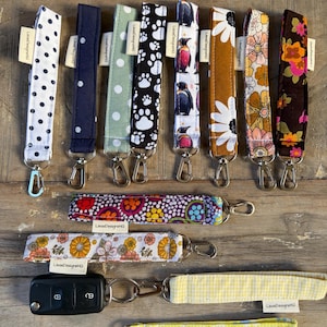 May include: A collection of fabric wristlet keychains featuring various patterns including polka dots, paw prints, penguins, daisies, and floral prints. Each strap includes a metal lobster clasp and a small white tag labelled LisasDesignsHQ.