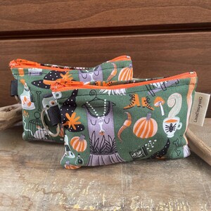 May include: Two small green zippered pouches with orange trim and black zipper pulls. The pouches are decorated with Halloween-themed illustrations, including pumpkins, witch hats, and dresses. The pouches are on a wooden surface.
