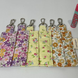Könnte beinhalten: Sechs Lippenbalsamhalter mit Blumenmuster und silbernen Schlüsselanhängern. Die Halter weisen verschiedene Muster auf, darunter rosa, lila, gelbe und orangefarbene Blumen. Ein rosa und blauer Lippenbalsam befindet sich neben den Haltern.