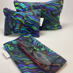 May include: Three zippered pouches with a green, blue and purple iridescent pattern. The pouches are made of fabric and have a zipper closure. The largest pouch is lying flat, the medium pouch is standing upright, and the smallest pouch is lying on its side. The pouches have a label that says "Remarkable Designs".