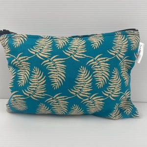 Può includere: Una pochette con cerniera blu turchese con un motivo a foglie beige. La pochette ha una cerniera nera e un'etichetta bianca che dice "Crafty Designs".