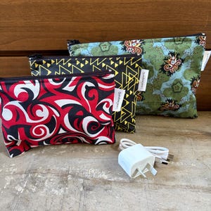 May include: Three rectangular zipped pouches in different fabric patterns: one with red, black, and white swirls, one with yellow geometric triangles on black, and one with green floral motifs. A white USB power adapter with a cable is in front.