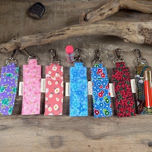 Può includere: Una collezione di portabalsamo per labbra in tessuto colorato con portachiavi argentati. I porta balsamo presentano vari motivi floreali nei toni dell'arancione, viola, rosa, rosso, blu e verde. Un tubetto di balsamo per labbra è visibile al centro.