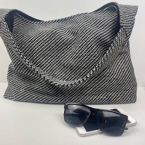 May include: A large black and white houndstooth tote bag with a black strap. Black sunglasses and two white smartphones are in front of the bag. The bag is made of a woven fabric.