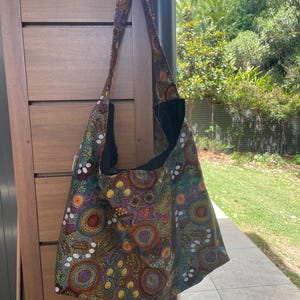 May include: A patterned shoulder bag in shades of brown, orange, yellow, and green. The bag features a repeating design of circles and dots. It has a long strap and a black interior. The bag is designed to be worn over the shoulder.