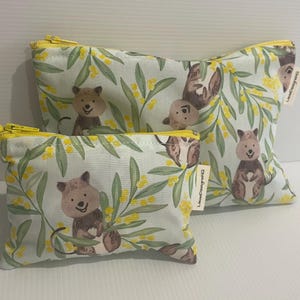 May include: Two zippered pouches featuring a pattern of cartoon quokkas and yellow floral accents on a pale green backdrop. The pouches have yellow zips and are in different sizes.