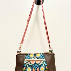 May include: A brown crossbody bag with a floral pattern on the front panel. The bag has a pink strap and gold-colored hardware. The floral pattern includes pink, yellow, and white flowers on a dark teal background.