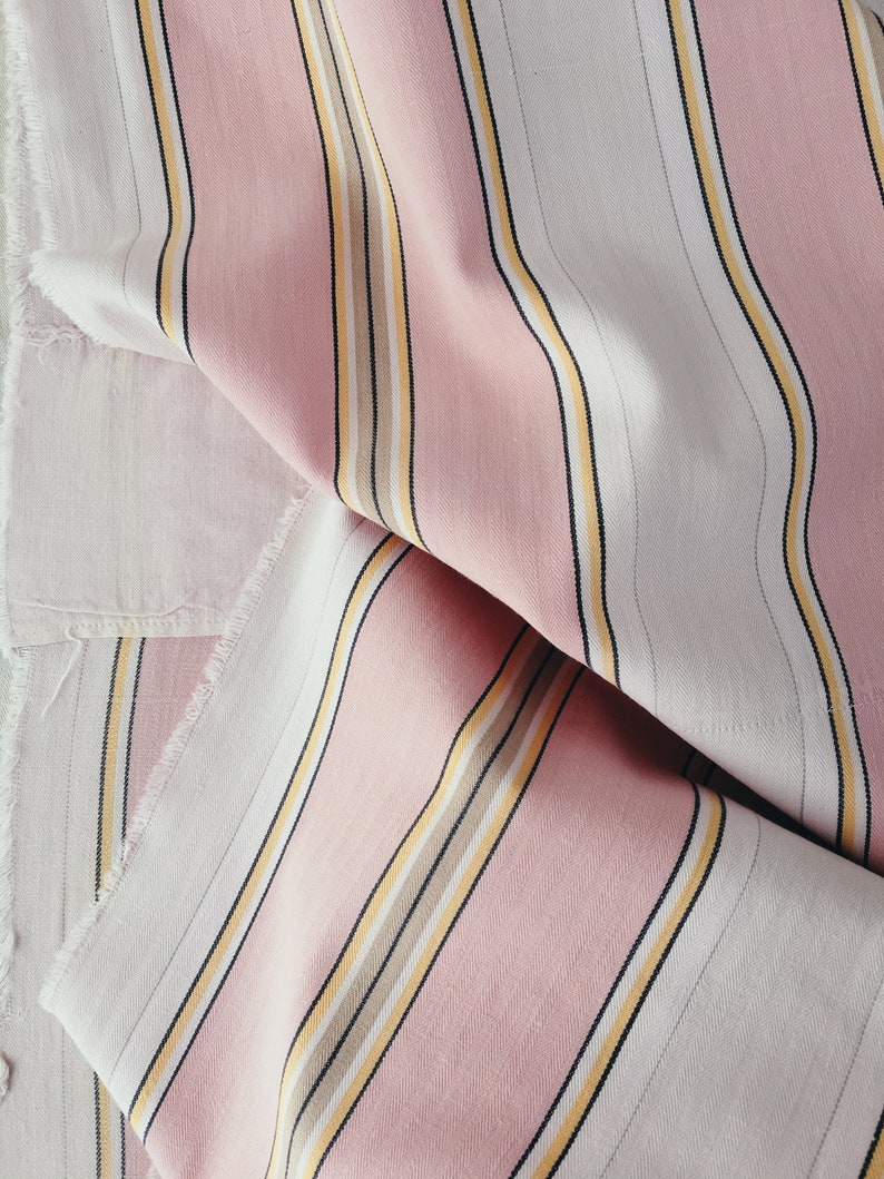 May include: Pink and white striped fabric with thin yellow and brown stripes. The fabric is folded and has a slightly frayed edge.