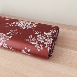May include: A roll of red fabric with a white floral pattern. The fabric is folded and lying on a light brown wooden surface.