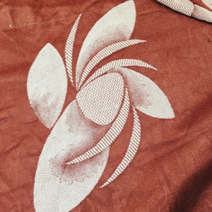 May include: A close-up of a rust-colored fabric with a white embroidered floral design. The design features a stylized flower with multiple petals.