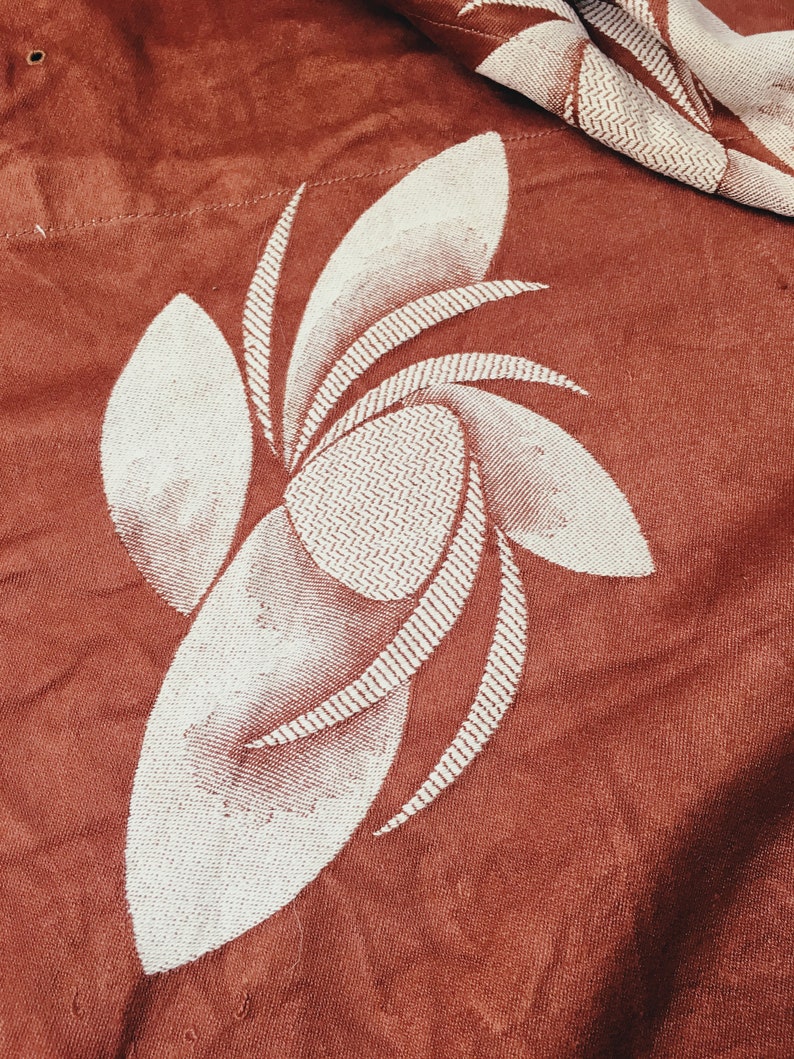 May include: A close-up of a rust-colored fabric with a white embroidered floral design. The design features a stylized flower with multiple petals.
