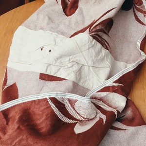May include: A brown and white woven throw blanket with a floral pattern. The blanket is folded and has a white patch sewn on one side.