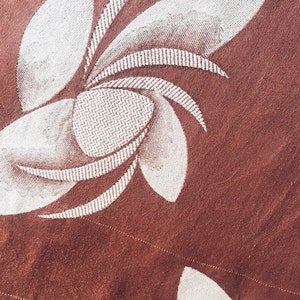 May include: A close-up of a brown fabric with a white embroidered floral design. The design features a stylized flower with multiple petals.