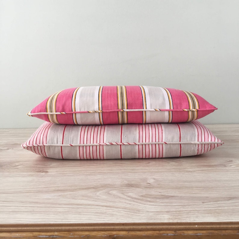 May include: Two striped pillows, one pink and white with yellow stripes and one gray and white with pink stripes. The pillows are stacked on top of each other on a wooden surface.