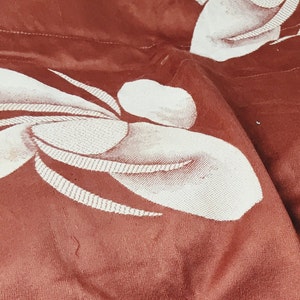May include: Close-up of a rust-colored fabric with a white embroidered floral pattern. The fabric is textured and has a subtle sheen.