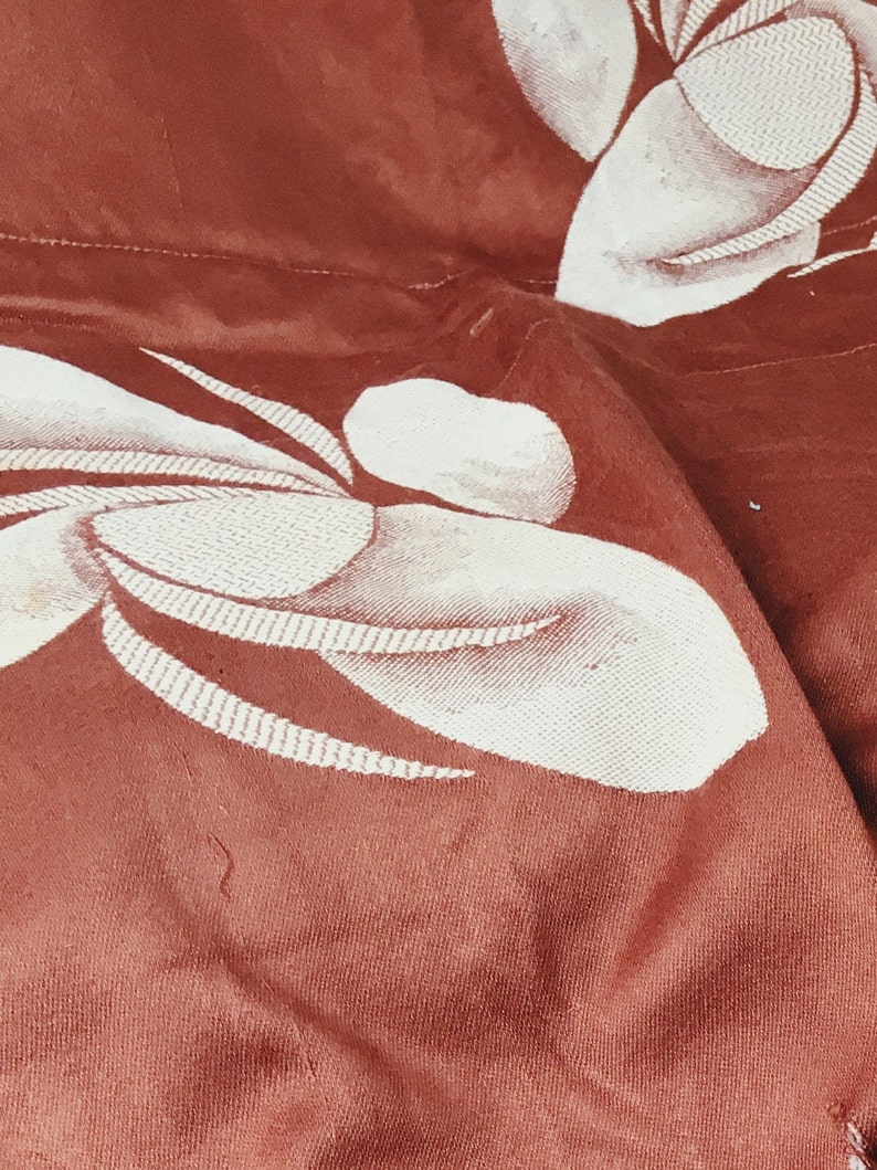 May include: Close-up of a rust-colored fabric with a white embroidered floral pattern. The fabric is textured and has a subtle sheen.
