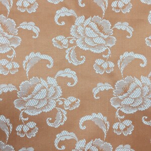 May include: Close-up of a peach-colored fabric with a repeating floral pattern. The design features large, white, stylized flowers and leaves with textured, striped details. The fabric has a subtle sheen, suggesting a satin or similar weave.