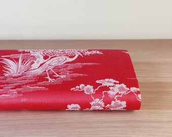May include: Red fabric with a white floral pattern featuring birds and branches. The fabric is folded in half and lying on a wooden surface.