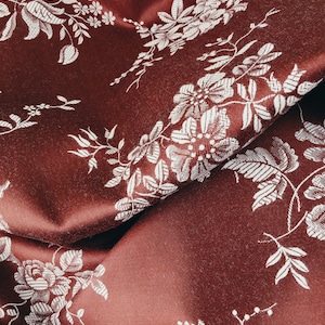 May include: Close-up of a red fabric with a white floral pattern. The fabric is textured and has a subtle sheen.