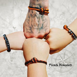 May include: Four wrists with matching bracelets in black and orange, and black and blue. The bracelets are knotted and some have white letter beads spelling out names. The text "Pirate Bracelets" is in the lower right corner.