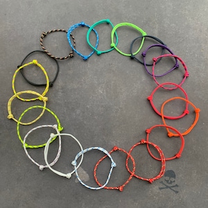 Peut inclure: Une collection de bracelets en corde colorés dans diverses teintes, dont noir, bleu, vert, jaune, blanc, rouge et orange. Chaque bracelet est noué et circulaire, avec un design minimaliste. Les bracelets sont disposés en cercle sur une surface gris foncé.